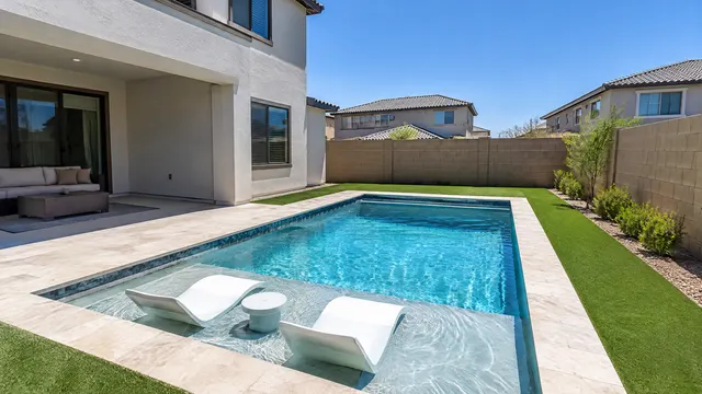 Brand-new compact rectangular pool with a sun shelf in the modest backyard of a recently built two-story home in a southeast Mesa master-planned community, small travertine deck, artificial turf, new block wall, rooftops of neighboring new homes, clear blue sky
