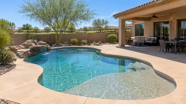 New freeform pool with a baja shelf behind a 2010s stucco home in east Mesa, Arizona, sandstone-color deck, covered patio, block wall with a green common area just visible beyond, palo verde tree, sunny day
