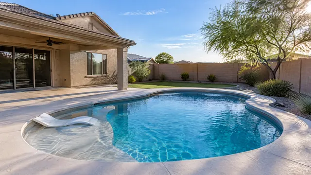 New kidney-shaped family pool with a short sun shelf behind a single-family stucco home in east Mesa, Arizona, light cool deck, small grass area, block wall, mesquite tree, sunny late afternoon