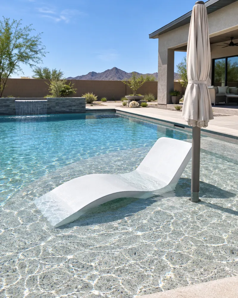 In-water lounge chair on a shallow baja shelf of a new pool, shade umbrella in a sleeve beside it, clear shallow water over light pebble finish