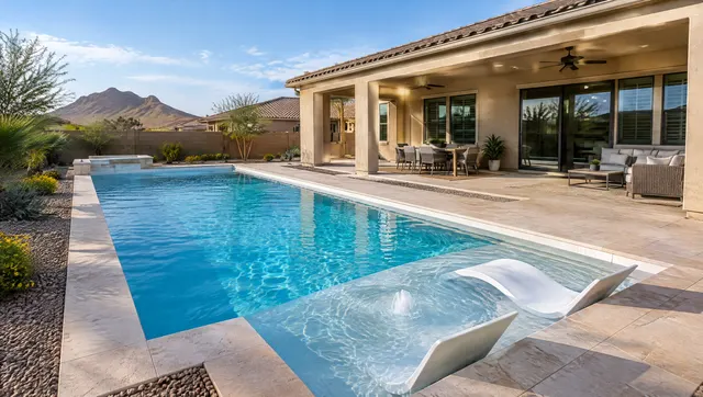 Newly built custom geometric swimming pool behind a single-story stucco Mesa home: long rectangle with a sun shelf at one end, clean white waterline tile, light sandstone coping, bright blue water, a covered patio with ceiling fans, crisp late-afternoon light