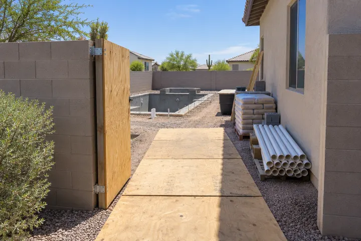 Pool construction access route in an Arizona side yard: temporary gate opening in a block wall, plywood protecting the ground, neat staging area with stacked materials, clean and organized