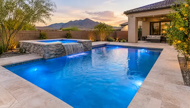 New raised spa with a sheer spillway pouring into a brand-new rectangular pool in an Arizona backyard at dusk, warm underwater lights glowing blue, stacked stone spa wall, travertine deck, citrus tree beside the patio